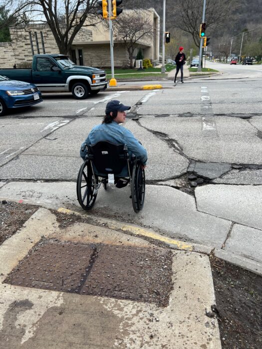 a person sitting in a wheelchair waits to cross a 4-lane road