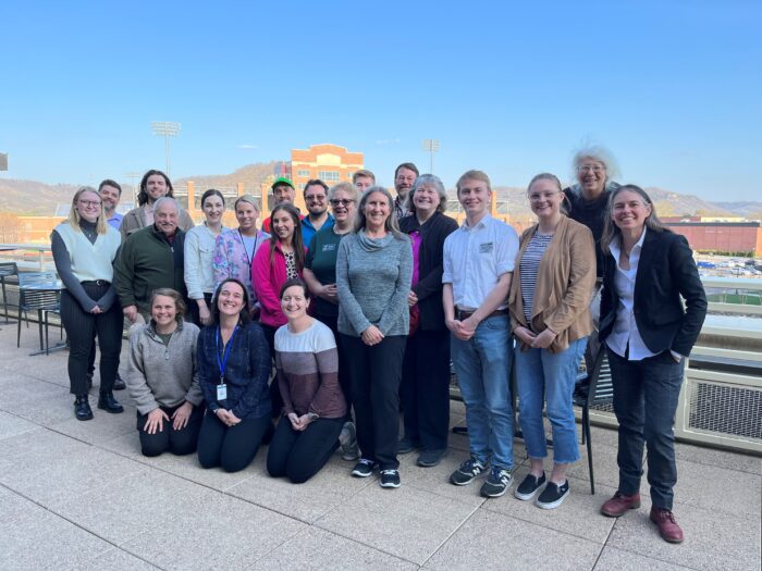 a group of smiling people gathered outside on a balcony with bluffs and blue sky in the background