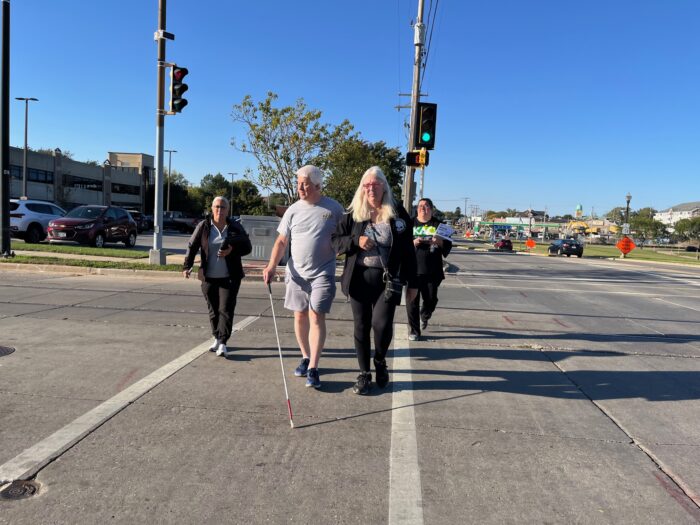 4 people cross a large road. one of them is using a white cane and holding the arm of th person next to him