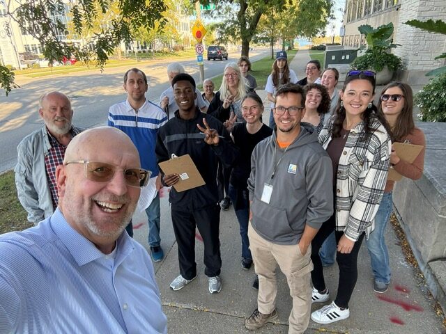 a group of 16 people smile for a selfie outside on a public sidewalk