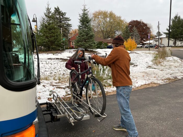 two people in front of a bus mounting a bicycle on a rack against a background of snow