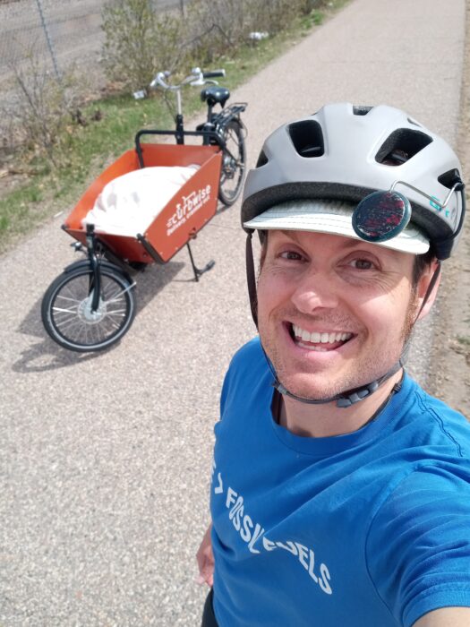 A white man wearing a blue t shirt and bike helmet is smiling in a selfie. A cargo bike is next to him on the asphalt.