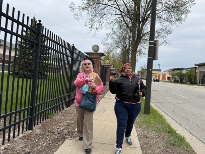 Two people on a sidewalk next to a tall black fence. One person is holding a speed gun.