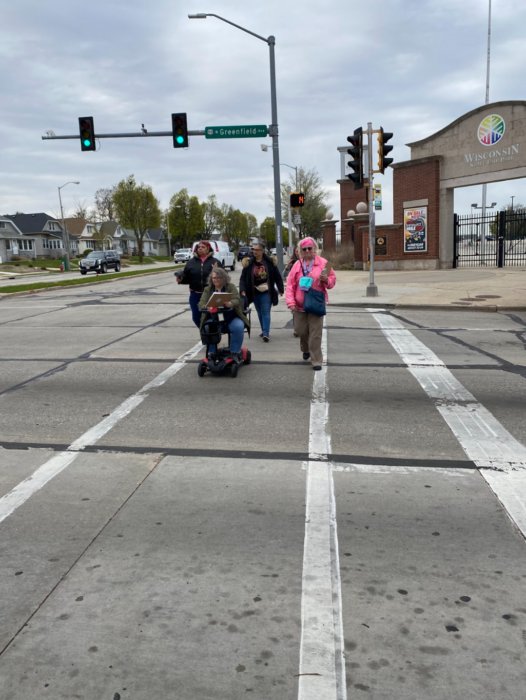 A group of people cross a wide street. One of them is on a motorized scooter.