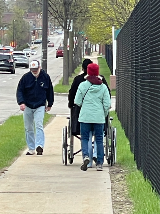 3 people walk down a sidewalk. One is facing the camera. The other two are walking away from the camera and pushing a wheelchair.