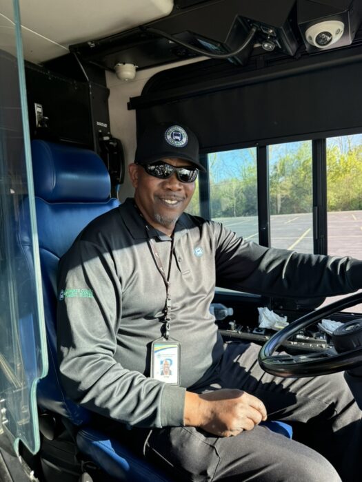 A smiling bus driver wearing dark sunglasses and an MCTS cap.