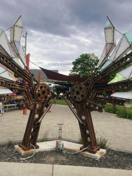 The image shows a public art installation in the shape of stretched out bird wings made out of various industrial parts