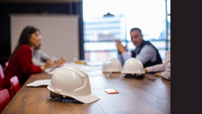 Image of individuals meeting to discuss construction or building project, with white hard hats on the wooden table