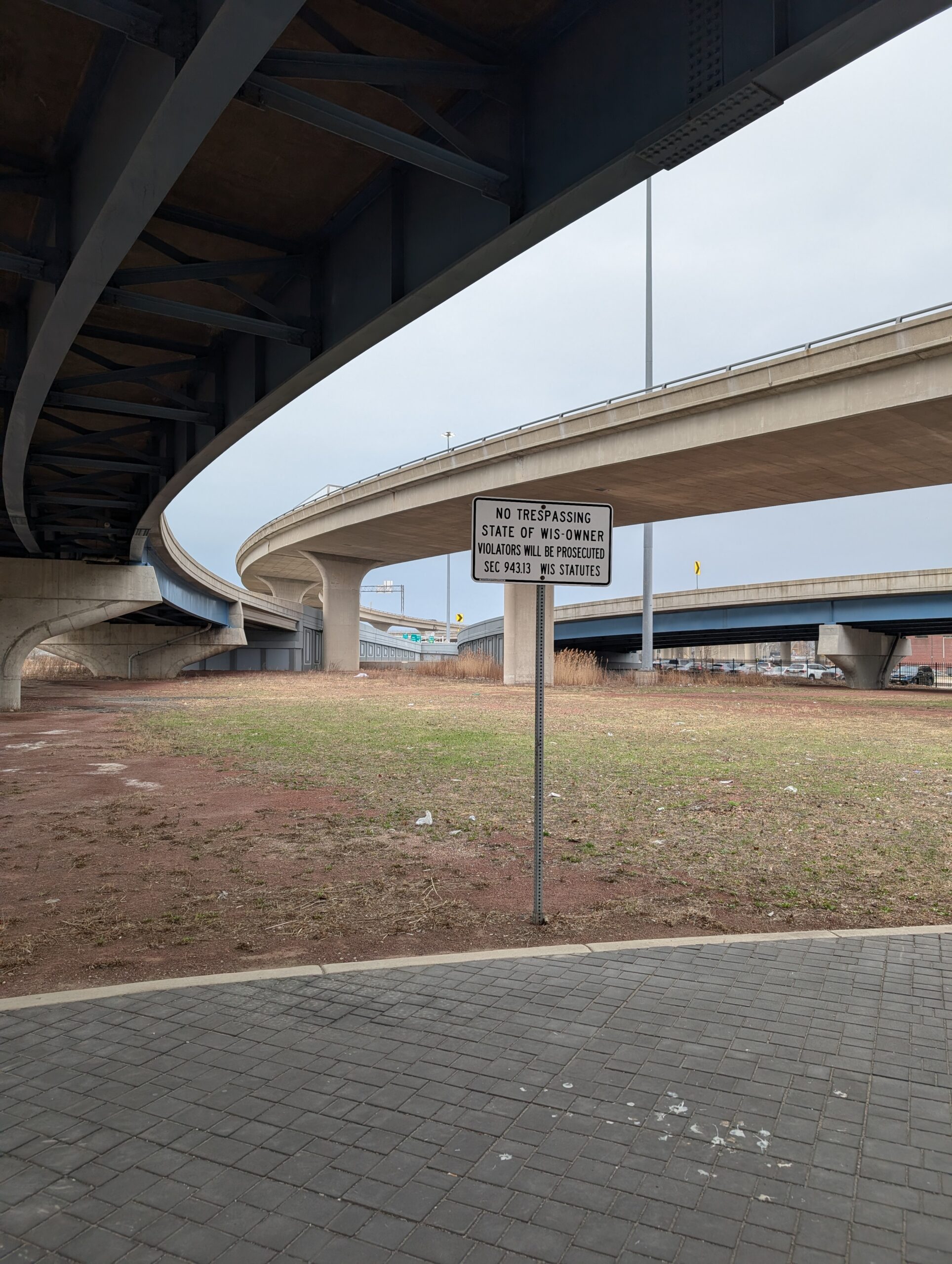 Image shows dead space under the I-794 Lake interchange ramps. A sign is posted stating "NO TRESPASSING STATE OF WIS-OWNER. VIOLATORS WILL BE PROSECUTED SEE 943.13WIS STATUES"
