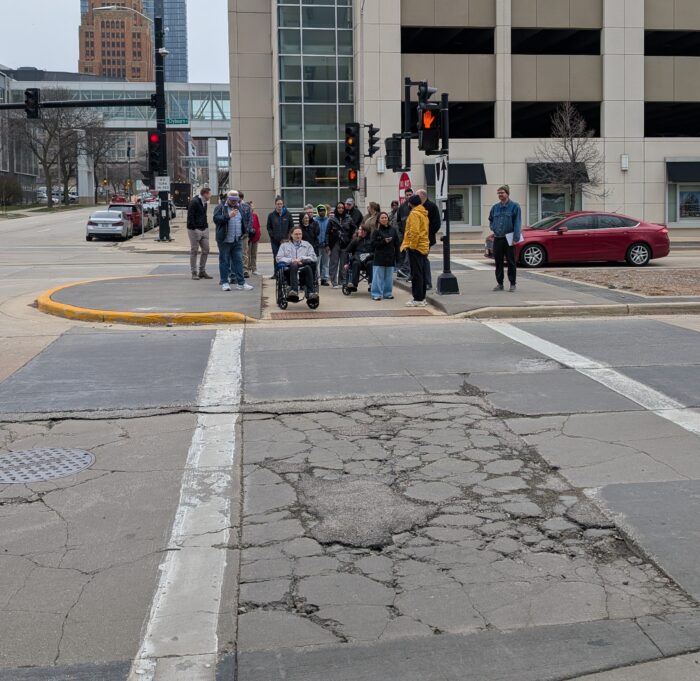The image depicts a group of participants waiting on the pedestrian refuge while crossing southward on the Van Buren St. and Clybourn Ave. intersection. A participant using a wheelchair is at the front of the crowd waiting to cross. Some of the group notice major cracks in the crosswalk.