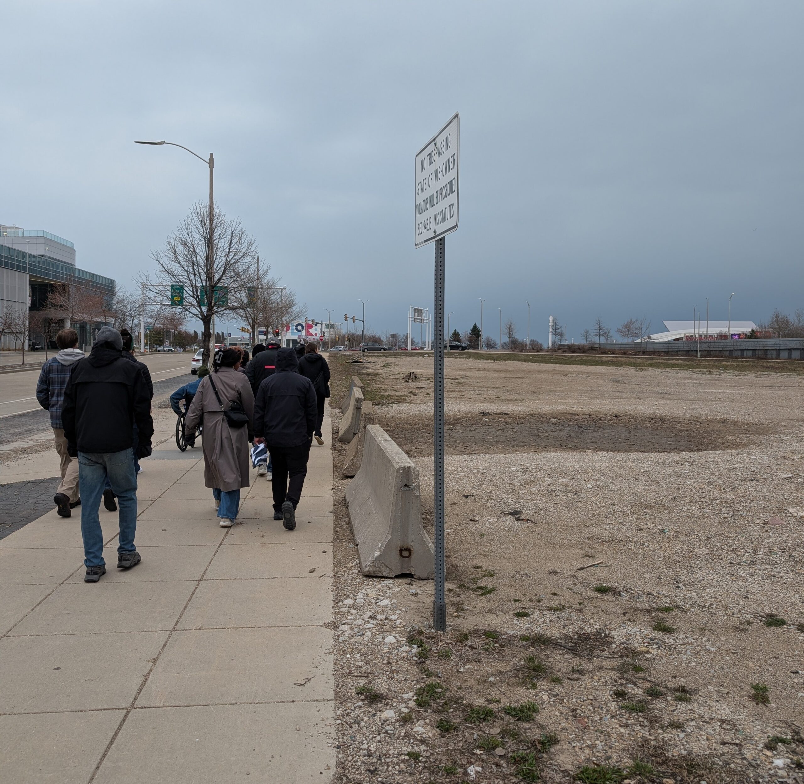 Image shows participants walking and rolling on the southside sidewalk of Clybourn Ave. adjacent to an open and empty dirt lot, toward the Lincoln Memorial Drive intersection. Discovery world is beyond the intersection in the horizon.